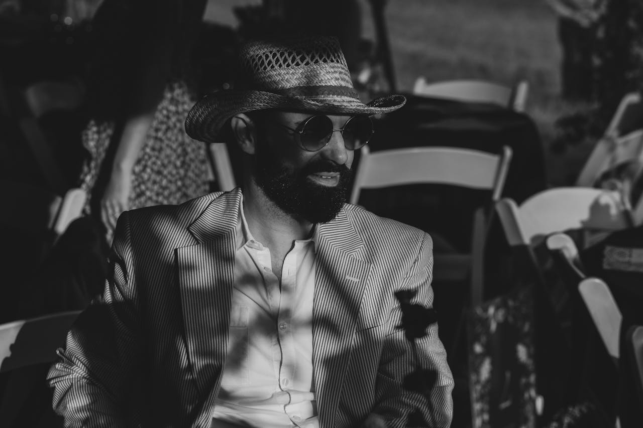 A man in a straw hat and striped blazer sits at an outdoor event with chairs around.